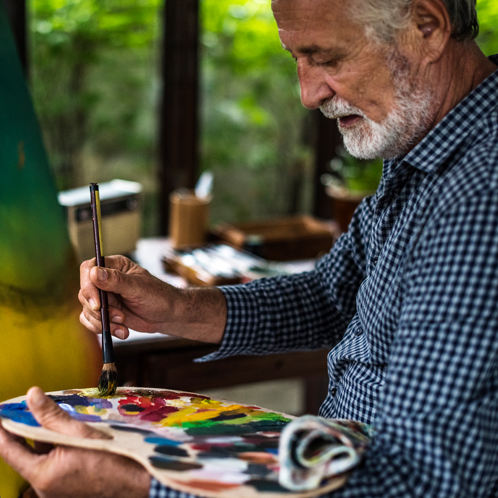Man with a white beard using an artist's palette and paintbrush with trees beyond the windows